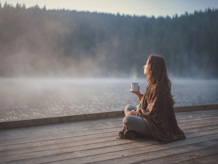 Woman meditating quietly by the lake with a warm drink